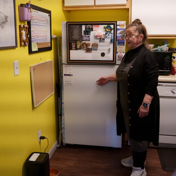 Woman standing in a yellow kitchen next to the refrigerator with notes and photos on it. A little box sits on the floor on the bottom left of the image.